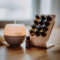 doTERRA Ultraschall-Diffuser mit warmem Licht und ein Holzständer mit mehreren ätherischen Ölen in dunklen Glasfläschchen auf einem Holztisch.